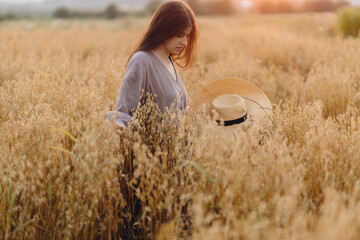 Stylish woman with straw hat standing in oat field in sunset light. Atmospheric tranquil moment....