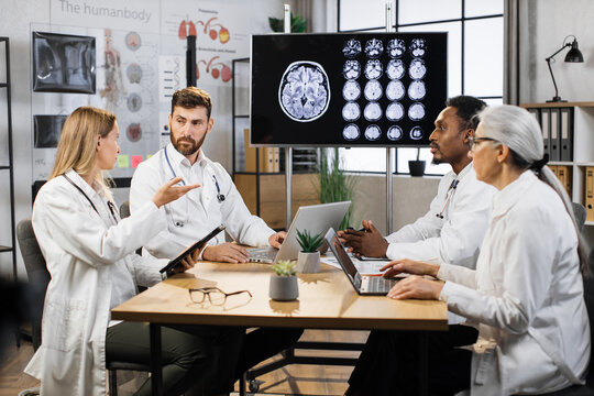 International medical team having meeting in conference room at modern hospital. Doctors discussing and interacting each other in laboratory with display showing brain MRI image on background.