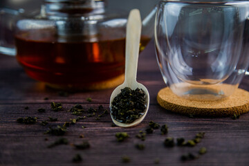 A wooden spoon with tea lies against a dark wood background, a mug and a teapot are visible from behind, a photo in dark colors. High quality photo