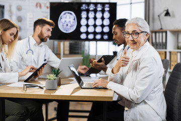 Portrait of senior female doctor sitting at desk with laptop and looking at camera. Group of multiethnic physicians taking online notes on different gadgets to patient history near.
