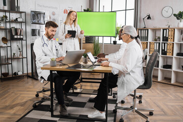 Caucasian female doctor ponting on monitor with green mock up screen while making presentation for international colleagues. Group of medical experts sitting at desk and listening briefing speech.
