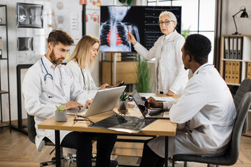 Qualified female doctor leading seminar to three multiracial interns at boardroom. Senior woman standing near digital screen and explaining results of chest x ray scan.