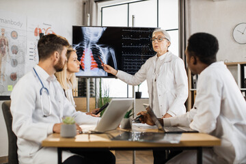 Group of multiethnic inters in lab coats sitting at desk with modern gadgets and listening attentively female head doctor. Senior woman pointing on x ray scan depicted on big monitor.