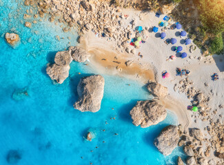Aerial view of blue sea, sandy beach with umbrellas, stones and rocks in water at sunset in summer....