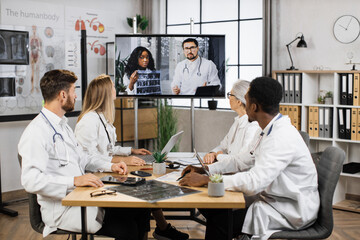 Fototapeta premium Group of four multicultural physicians watching online lecture about MRI diagnosis in modern hospital. Confident doctors analyzing ways of successful treatment during video conference with colleagues.