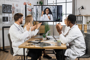 Team of multicultural doctors in white lab coats clapping hands during online video conference after successful speech of excited female scientist. Focus on african american woman on monitor screen.