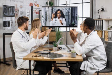 Fototapeta premium Team of multicultural doctors in white lab coats clapping hands during online video conference after successful speech of excited female scientist. Focus on african american woman on monitor screen.
