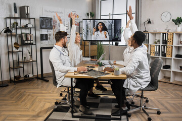 Group of motivated international scientists sitting at table and raising hands in hospital conference room. Excited multiethnic workers unanimously voting for suggested idea during online video call.