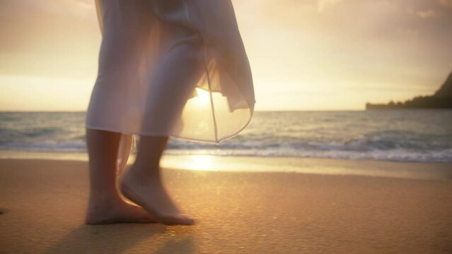 Close up girl in romantic white boho style wedding dress walking barefoot along ocean waves on paradise sandy beach at beautiful golden glowing sunset on Hawaii island. Slow motion nature RED 6K shot