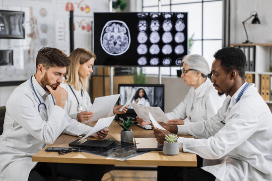Multicultural group of four medical researchers having online training in laboratory with monitor on background showing brain MRI image. Collegues sharing ideas while holding patient history list.
