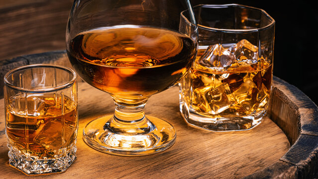 Glass of whiskey with ice, glass of cognac, glass of bourbon with ice stand on wooden barrel of whiskey against background of wooden background. Alcoholic beverages whiskey, cognac, brandy close-up.