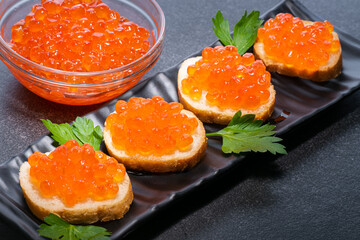 Caviar on loaf of butter on black rectangular plate. Next to butter rolls are green edible herbs. In upper left corner there is transparent plate with red caviar.