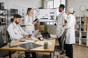 Group of multiethnic concentrated doctors standing near screen during working sesion in lab boardroom. Caucasian male working on laptop while team of colleagues talking about genetic engineering.