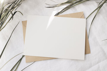 Summer wedding stationery mock-up, desk scene. Blank horizontal greeting card, dry green palm leaf. In sunlight. White linen table background. Tropical styled photo, web banner. Flat lay, top view.