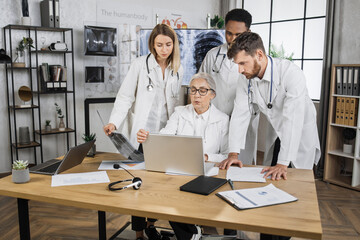 International team of three interns having urgent meeting with senior female doctor in hospital conference room. Workerks in white lab coats using modern laptop for analysing patients medical record.