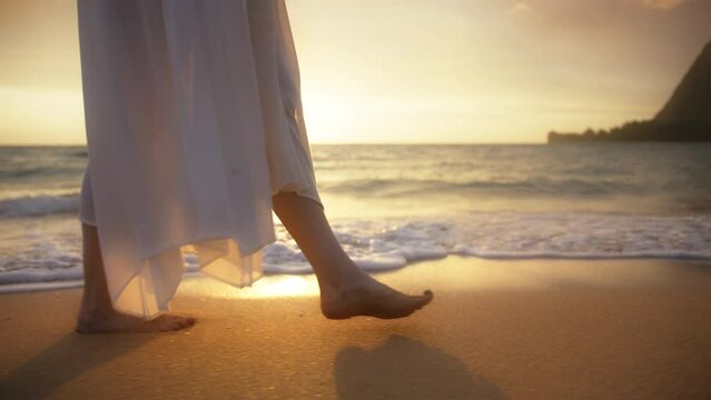Slow motion woman feet walking barefoot by beach at golden sunset leaving footprints in sand. Female tourist on summer vacation in Kauaii, Hawaii island USA. Woman in beautiful waving dress at sunset