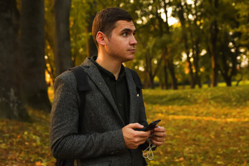 Defocus young man holding phone. Man using smart phone in autumn park. Typing text message or reading social media at mobile phone. Fall background. Cell phone. Backpack. Outside. Out of focus