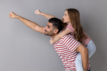 Cheerful daddy piggybacking daughter, making flying superhero hand, fist forward gesture, little...