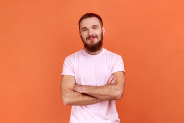 Portrait of bearded man standing with crossed arms, looking at camera, expressing positive emotions, being in good mood, wearing pink T-shirt. Indoor studio shot isolated on orange background.