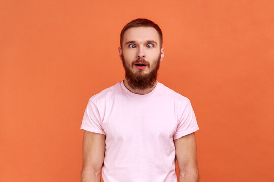 Portrait of bearded man looking cross-eyed with stupid smile, fooling around, making silly face, brainless comical expression, wearing pink T-shirt. Indoor studio shot isolated on orange background.