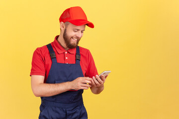 Portrait of satisfied smiling courier or craftsman standing with with smartphone in hands, taking online order, being ready to work. Indoor studio shot isolated on yellow background.