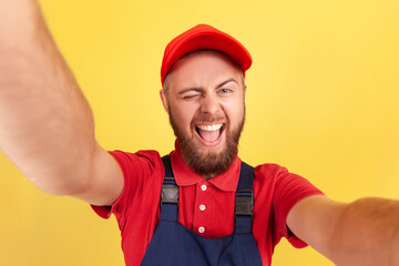 Portrait of funny playful bearded worker man wearing uniform taking selfie, looking and winking at camera POV, point of view of photo. Indoor studio shot isolated on yellow background.