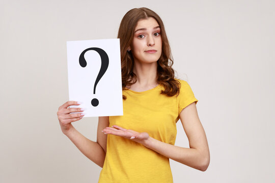 Young attractive brown haired young woman in yellow casual style T-shirt holding paper with question mark, making decision, solving problem. Indoor studio shot isolated on gray background.