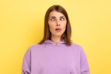 Portrait of young adult foolish woman with hair standing with crossed eyes and tongue out, childish behavior, wearing purple hoodie. Indoor studio shot isolated on yellow background.
