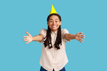 Come into my arms. Friendly woman with black dreadlocks and party cone on head giving free hugs with outstretched hands, welcoming inviting to embrace. Indoor studio shot isolated on blue background.