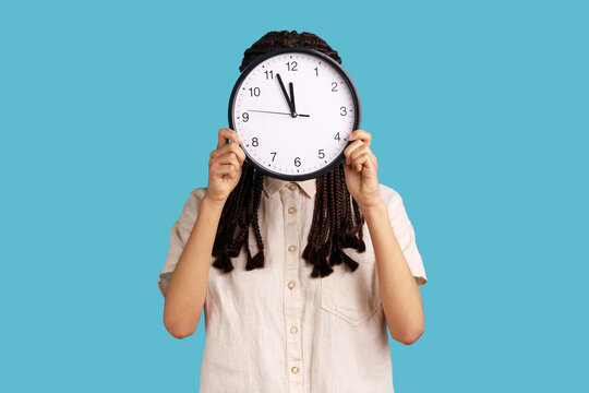 Time to work. Woman with dreadlocks hiding face behind big wall clock, time management, schedule and business meeting appointment, wearing white shirt. Indoor studio shot isolated on blue background.