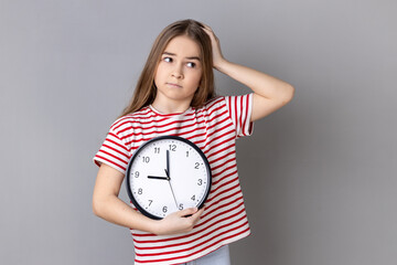 Portrait of confused uncertain little girl wearing striped T-shirt holding big wall clock in hands,...
