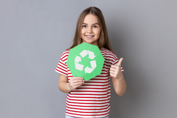 Portrait of little girl wearing striped T-shirt holding in hands green recycling sing, ecology concept, looking at camera and showing thumb up. Indoor studio shot isolated on gray background.