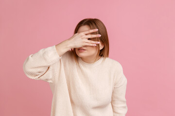 Blond woman covering eyes with hand, peeking through fingers and feeling shy curious about secret, watching forbidden content, wearing white sweater. Indoor studio shot isolated on pink background.