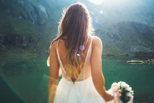 Back view of a bride with brunette hair in a white long wedding dress standing on the edge holding a wedding bouquet looking at the mountain lake. Lifestyle