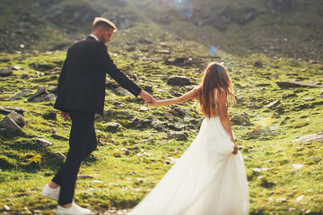 Naklejka na ściany i meble Bride in a dress with lace and groom holding hands and walking during their travel through the mountains. Nature landscape.