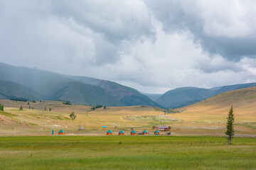 Scenic view from sunlit grassy steppe to small houses on background of green high mountain range in rainy clouds. Dramatic landscape with meadow and small lodges in mountains at changeable weather.