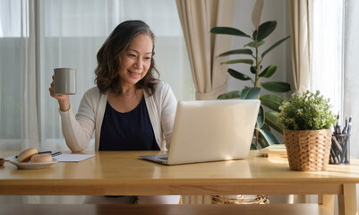 Smiling elegant senior woman drinking coffee and spending time with technology device in living room.