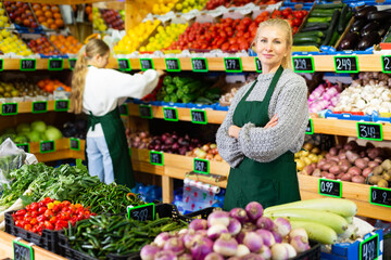 Portrait of smiling confident saleswoman of greengrocery store standing among shelves with fresh fruits and vegetables