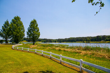 a gorgeous summer landscape at Lake Acworth with rippling blue lake water surrounded by lush green grass and trees with a white wooden fence and a gorgeous clear blue sky at Cauble Park in Acworth