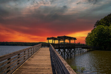 Fototapeta na wymiar a long brown wooden bridge over the rippling blue waters of Lake Acworth surrounded by lush green trees and plants with powerful clouds at sunset at Cauble Park in Acworth Georgia USA