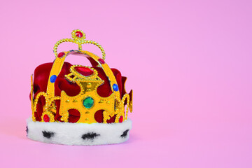 Toy plastic royal crown decorated with fake gems, fur and red cloth on pink background, studio...