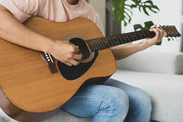 Young man is sitting on sofa at home next to the window with tree and playing the acoustic guitar on vacation. He plays guitar as a hobby and he feels relaxed and happy when he hears the music.