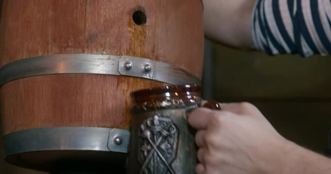 Person pours a drink into a ceramic mug with emblem from a hole in wooden barrel, close up. Storage of alcohol in the wine cellar. Authentic themed pirate bar