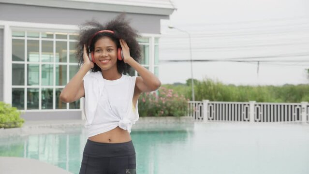 Young African American woman wearing headphones listening music standing dancing, female wearing earphones while enjoy with happiness at poolside, emotion and feeling, lifestyles concept.