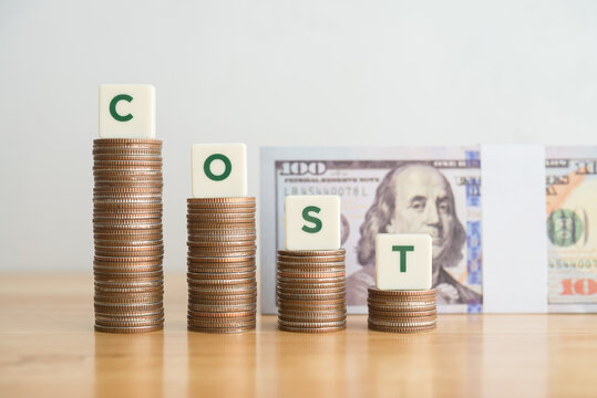 Word COST on stacks coin as graph chart falling down and US dollar banknotes background on wooden table. Concept of cost reduction, cost cut in business or production, optimize continuous improvement.