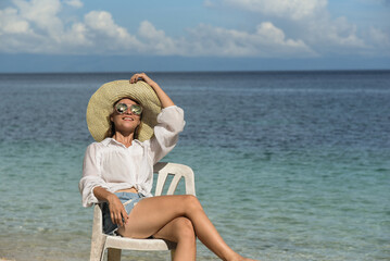 A lady in a straw hat, sunglasses, white shirt and denim shorts is sitting on a chair. A young girl is relaxing on the beach. Travel, happy holidays, tourism, Philippines, summer season, copy space