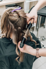 Fototapeta premium Hair stylist prepares woman makes curls hairstyle with curling iron. Long light brown natural hair