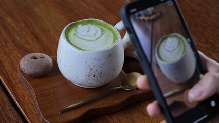 On the table is a mug with a hot drink with green foam, which is photographed on a smartphone by a blogger for their social networks. A food blogger takes a photo of the macha drink for his blog. - obrazy, fototapety, plakaty