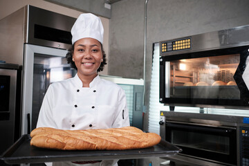 Portrait of African American female chef in white cooking uniform looking at camera with cheerful smile and proud with tray of baguette in kitchen, pastry foods professional, fresh bakery occupation. - Powered by Adobe