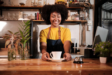 Fototapeta na wymiar African American female barista offers cup of coffee to customer with cheerful smile, happy service works in casual restaurant cafe, young small business startup entrepreneur.